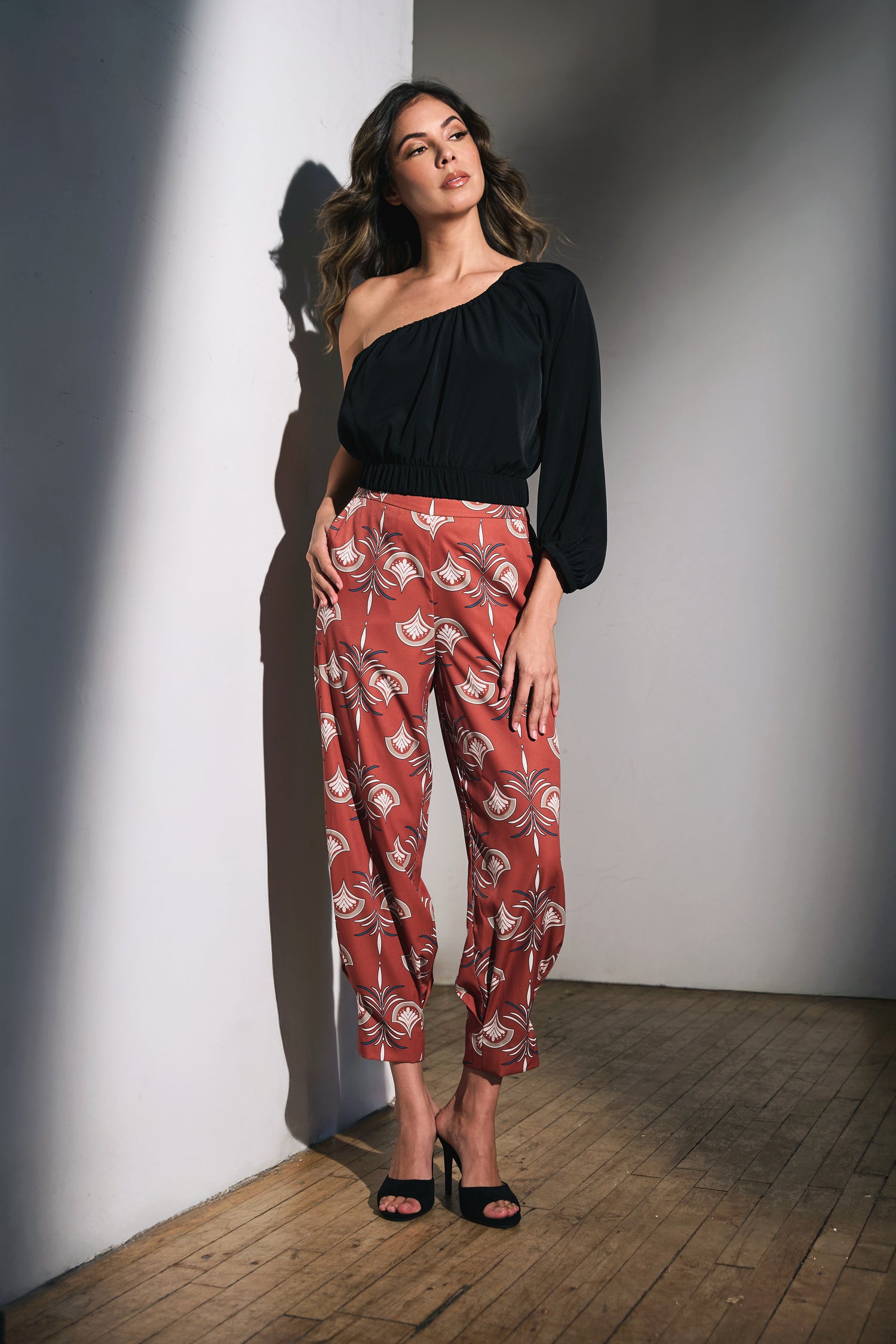 Woman wearing a Lorrel black top and bottom,  patterned pants standing against a gray studio wall.